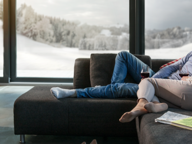 Personen sitzen entspannt auf einem modernen, dunklen Sofa vor einem knisternden Kaminfeuer, mit Blick auf eine verschneite Landschaft durch grosse Panoramafenster. Ein Korb mit Holz steht neben dem Kamin, Zeitschriften und Schuhe liegen im Wohnbereich.